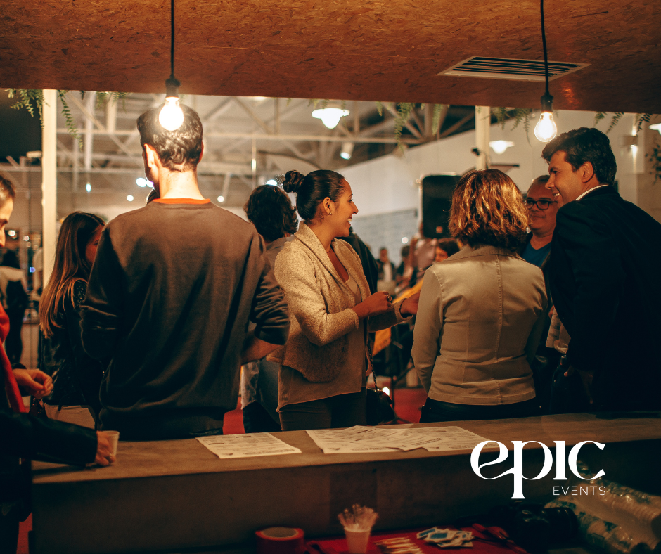 A group of diverse professionals networking at an indoor event with warm lighting, sharing conversation around a high-top counter with event materials.