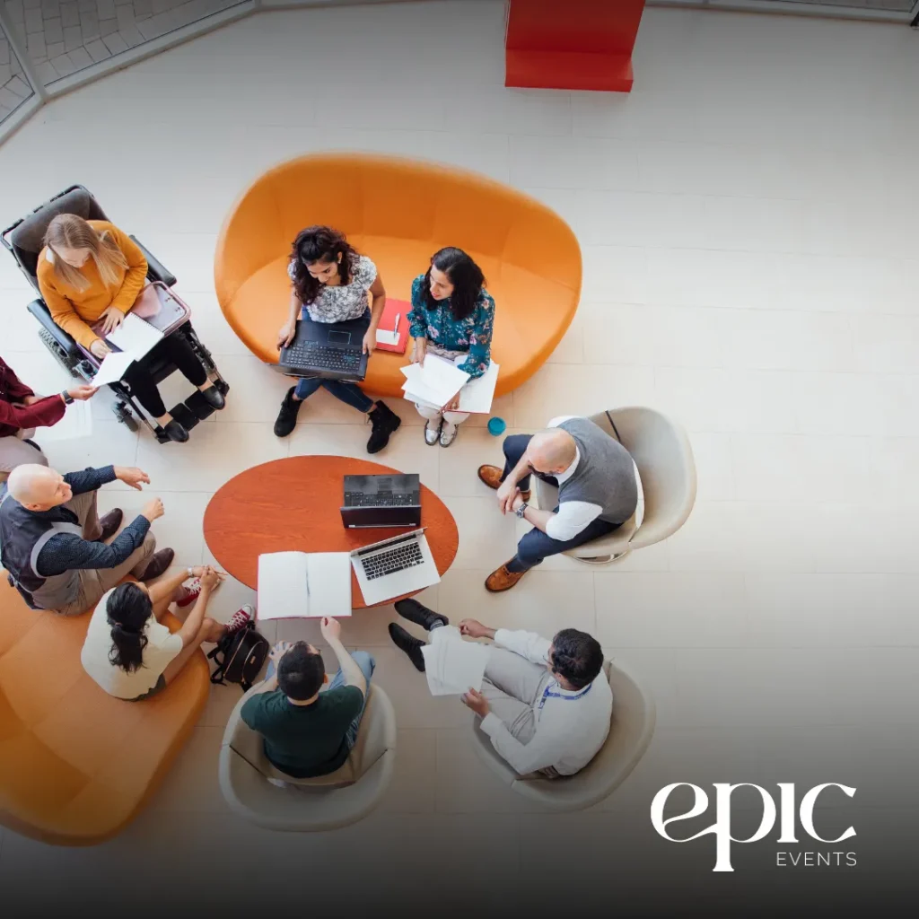 Group collaboration in a modern setting, featuring diverse individuals seated around a circular table with laptops and documents, emphasizing effective communication and teamwork in event logistics planning.